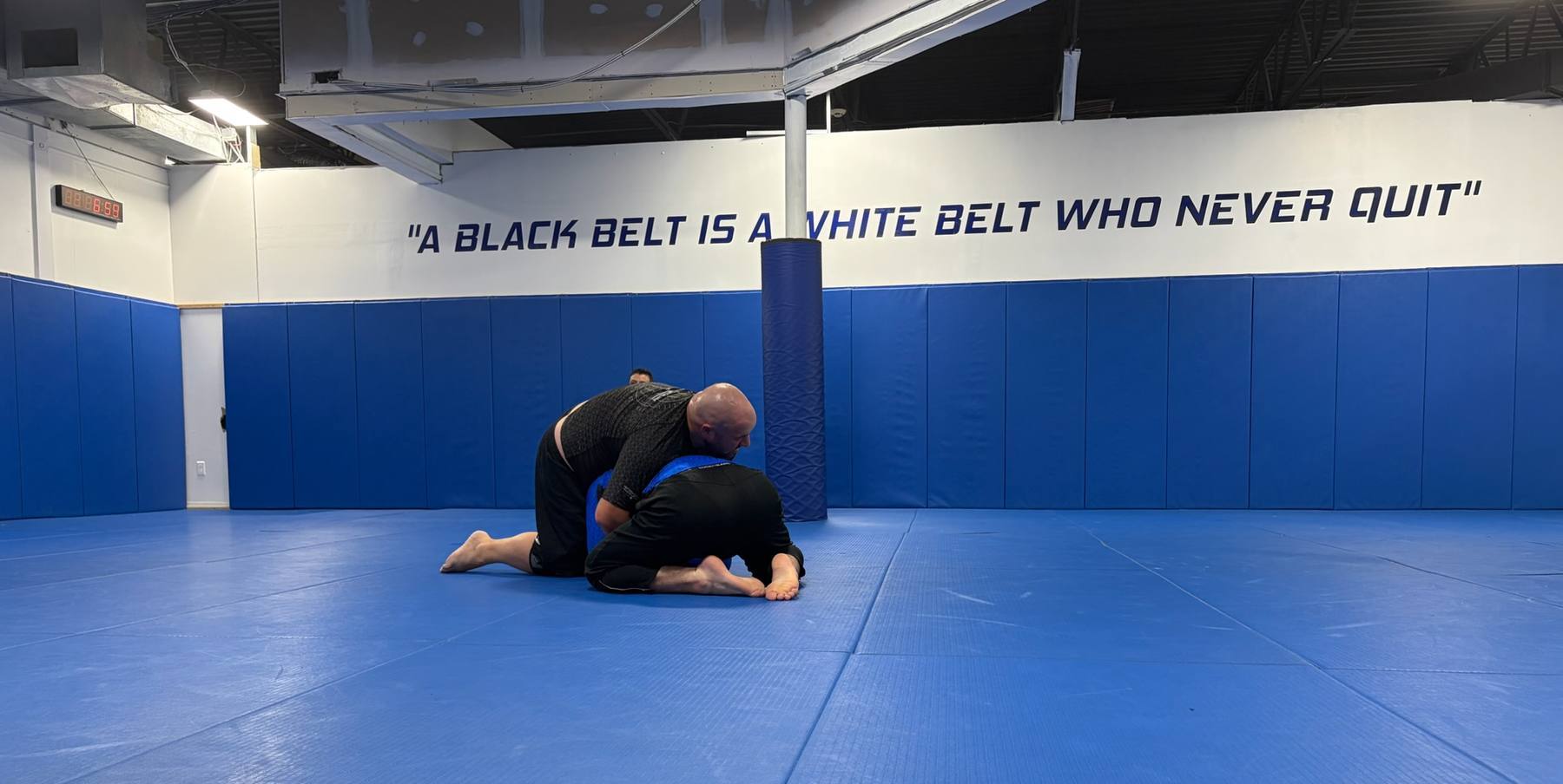 Two students grappling on the blue mat under the academy wall slogan